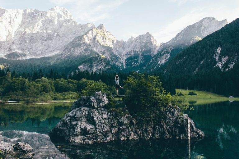 Eine Frau steht für ein Yoga Retreat in den Bergen auf einem Fels mitten im See im Yoga Baum